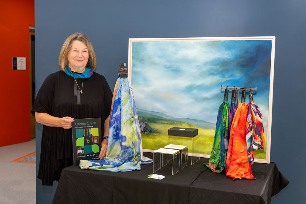 Artist Hazel Green with one of her paintings in the background and some silk scarves to the forefront.